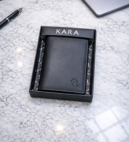 Black leather wallet in a KARA box on a marble surface with a keyboard and laptop in the background.