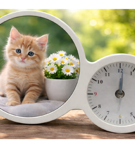 White clock with a round window showing a kitten and flowers on a wooden surface with a blurred green background